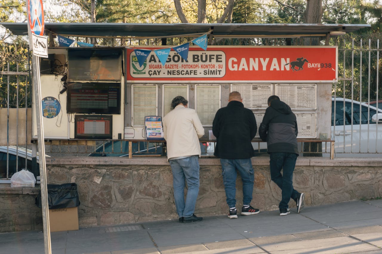 Three men at a street kiosk in Turkey, checking sports betting results on a sunny day.